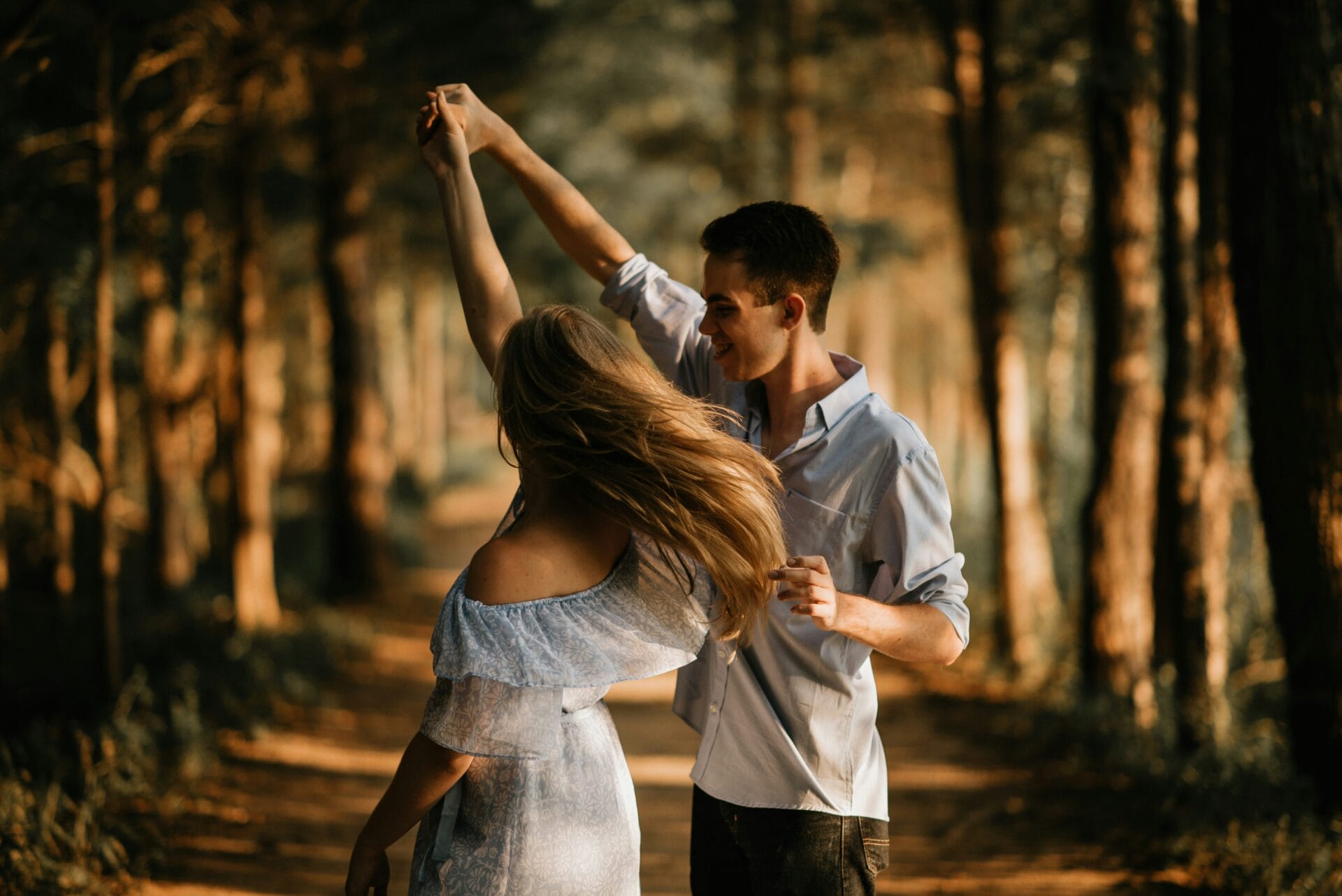 Un couple danse sur un sentier forestier, la lumière du soleil filtrant à travers les arbres, capturant l'essence des relations et des rencontres où la connexion s'épanouit et les moments de joie illuminent leur lien.