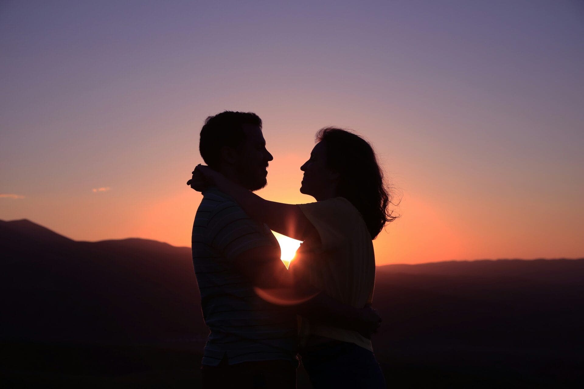 Silhouette d'un couple enlacé avec en arrière-plan un coucher de soleil sur un paysage montagneux.