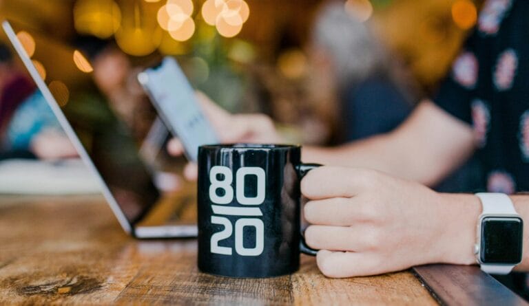 Une personne sirote une tasse noire avec l'inscription " 80/20 ", symbolisant le principe de Pareto dans les relations, alors qu'elle est assise devant un ordinateur portable ouvert et un smartphone sur une table en bois.