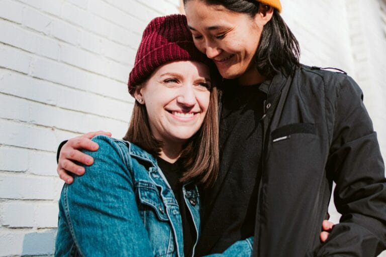 Deux personnes se tiennent l'une à côté de l'autre, souriant et s'étreignant devant un mur de briques blanches. Elles portent toutes deux un bonnet et une veste décontractée. Leur étreinte incarne l'essence des langages de l'amour, renforçant leur relation par la chaleur et la compréhension.