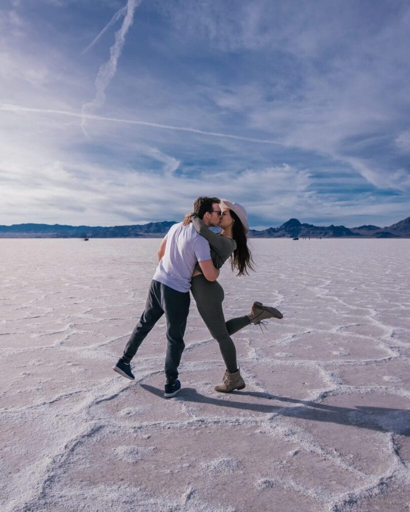 Un couple partage un moment candide, s'embrassant au milieu d'un vaste désert de sel sous un ciel partiellement nuageux, avec des montagnes visibles au loin - une idée de pose parfaite pour une séance photo de couple inoubliable.