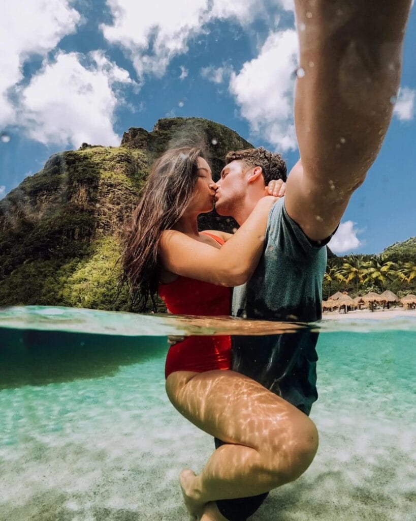 Un couple partage un moment candide, s'embrassant à genoux dans une eau claire et turquoise. La vue partagée capture leur bonheur au-dessus et au-dessous de la ligne de flottaison, sur fond de verdure tropicale luxuriante et de ciel bleu, une idée de pose parfaite pour une séance photo romantique.