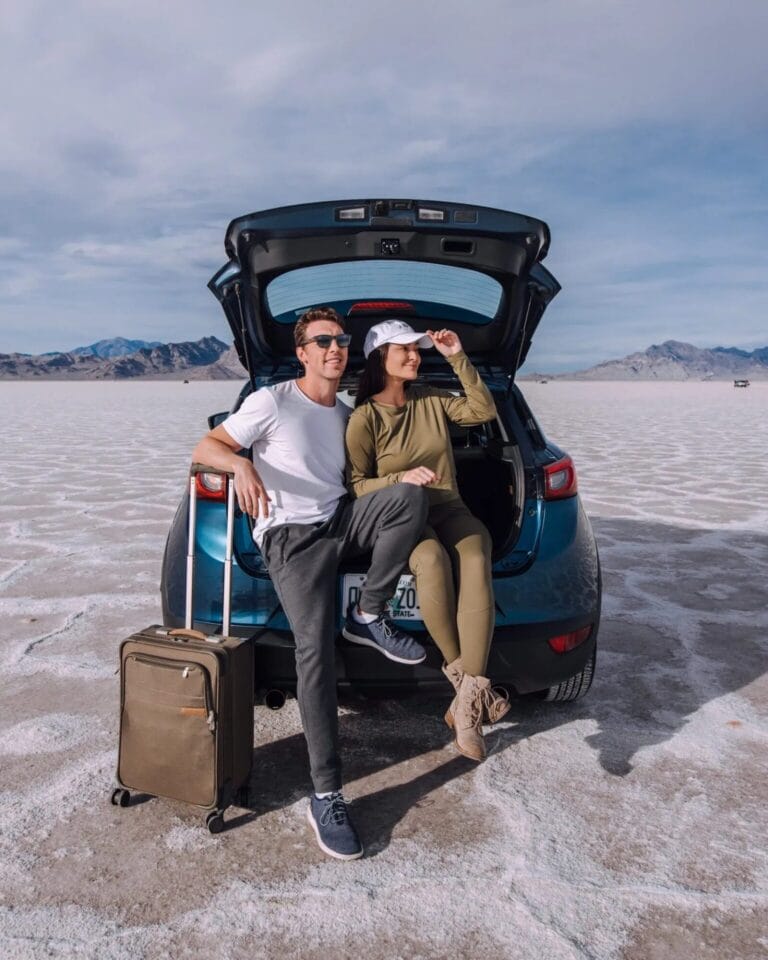 Un homme et une femme profitent d'un moment de spontanéité assis sur le coffre ouvert d'une voiture garée dans un désert de sel. Habillés de manière décontractée et portant des lunettes de soleil, ils trouvent des idées de pose pour leur séance photo de couple, avec une valise à côté d'eux et des montagnes en arrière-plan.