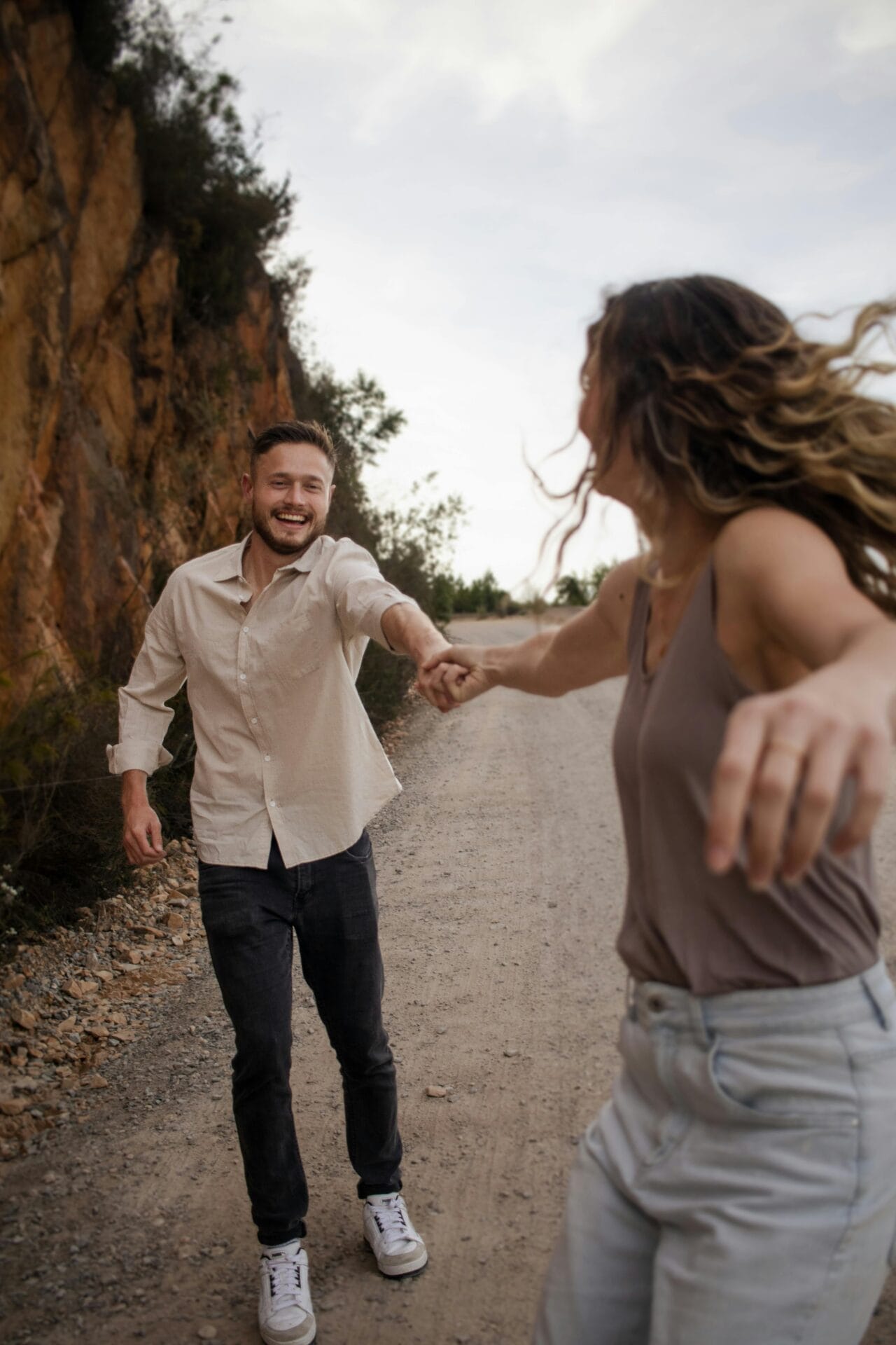 Un homme Gémeaux vêtu d'une chemise beige et d'un jean foncé tient joyeusement la main d'une autre personne vêtue d'un débardeur marron et d'un jean clair, en marchant sur un chemin de terre à côté d'une falaise rocheuse. La scène transmet le bonheur et le mouvement, capturant l'essence de l'aventure amoureuse.