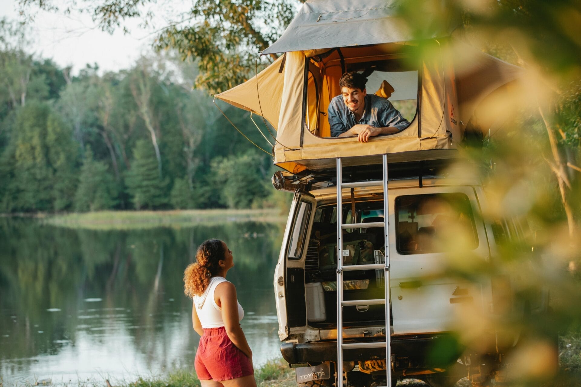 Une femme se tient au bord d'un lac, regardant son homme Gémeaux qui sourit depuis une tente sur le toit de leur véhicule. La forêt verte et luxuriante et l'eau calme en arrière-plan suggèrent une expérience de camping sereine qui capture la spontanéité et le charme d'une rencontre avec un Gémeaux.