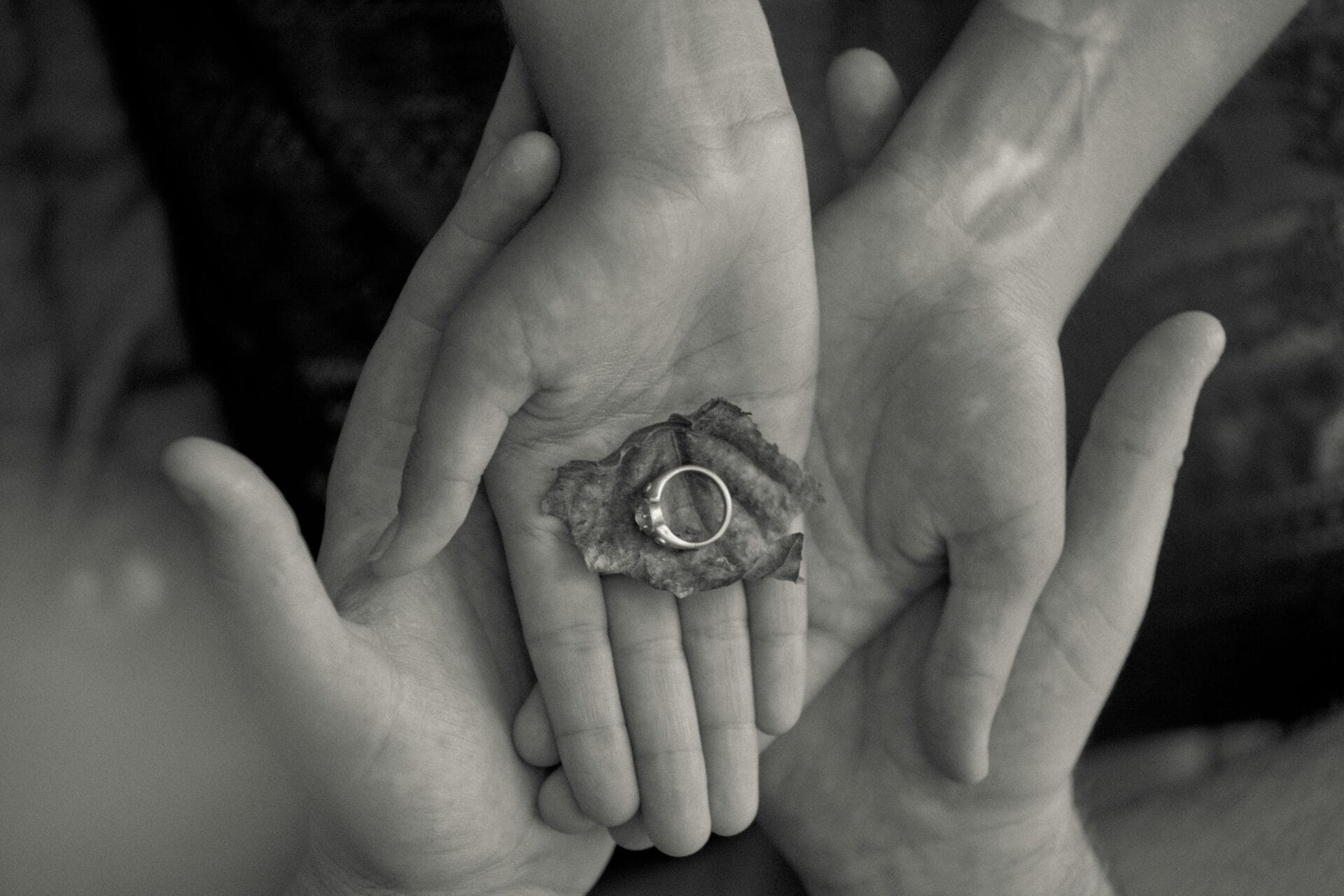 Quatre mains bercent délicatement une feuille sèche sur laquelle est placée une bague, créant ainsi un sentiment d'unité et d'attention. Cette métaphore visuelle de la compatibilité est capturée en noir et blanc, soulignant les textures et les contours des mains et de la feuille, tout comme la compréhension de la nature inébranlable d'un homme Taureau dans les relations amoureuses.