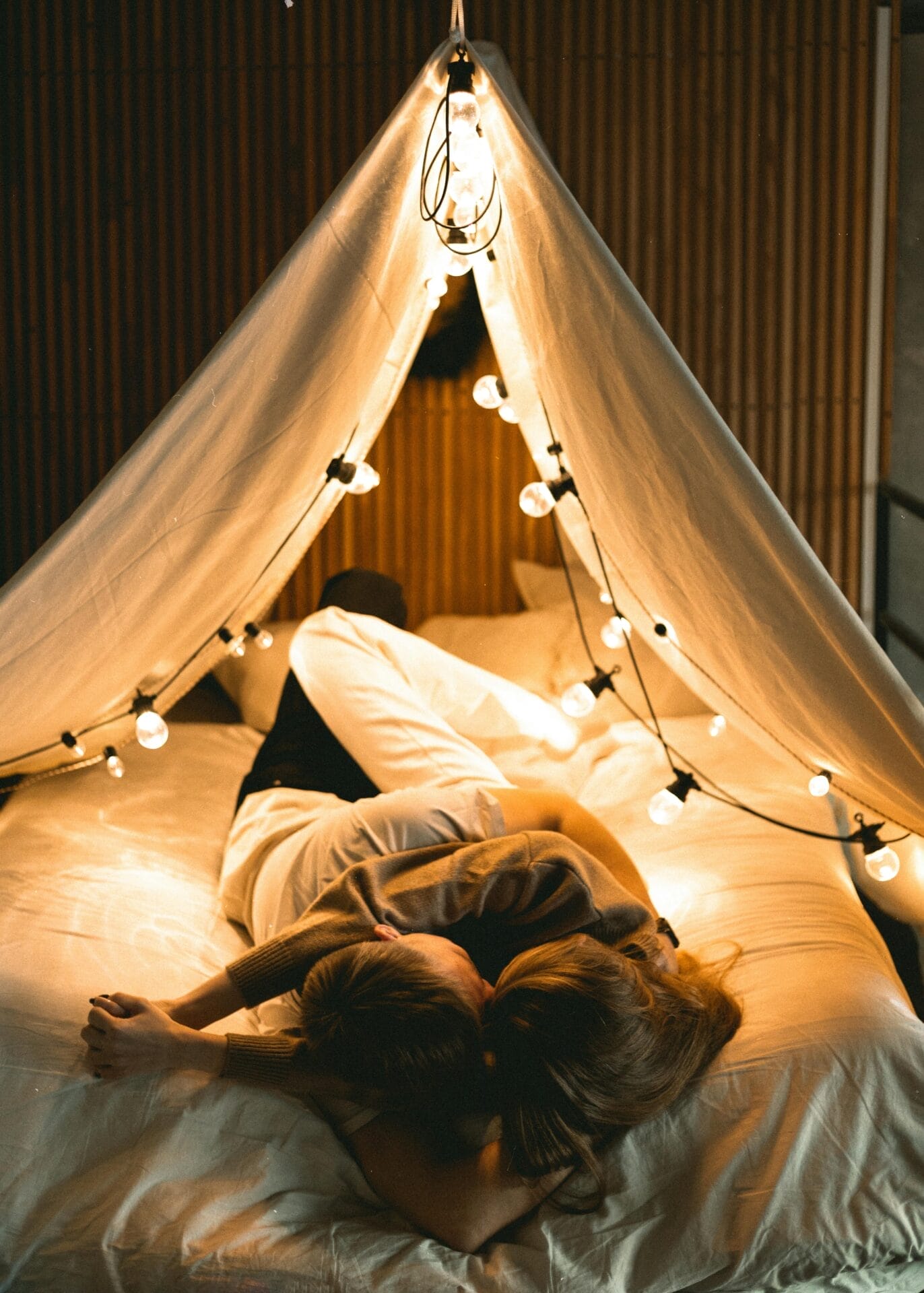 Un couple est allongé sur un lit sous un baldaquin confortable, semblable à une tente, orné de lumières féeriques. La lueur chaude et ambiante crée une atmosphère intime et détendue, parfaite pour explorer la compatibilité. Cette retraite à la lumière tamisée est idéale pour des conversations profondes et des connexions significatives.