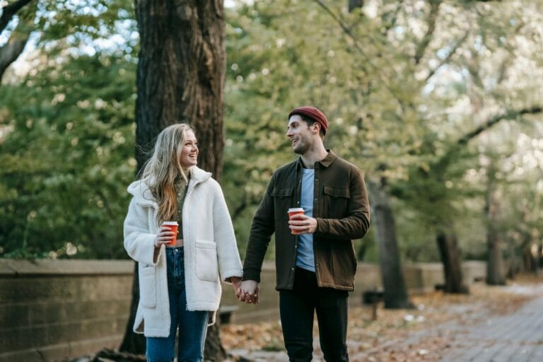 Un couple souriant marche main dans la main le long d'un chemin bordé d'arbres, tenant des tasses de café rouges. Ils semblent détendus et heureux, profitant d'une expérience de rencontre sobre et populaire au milieu du feuillage vert et doré de l'automne.