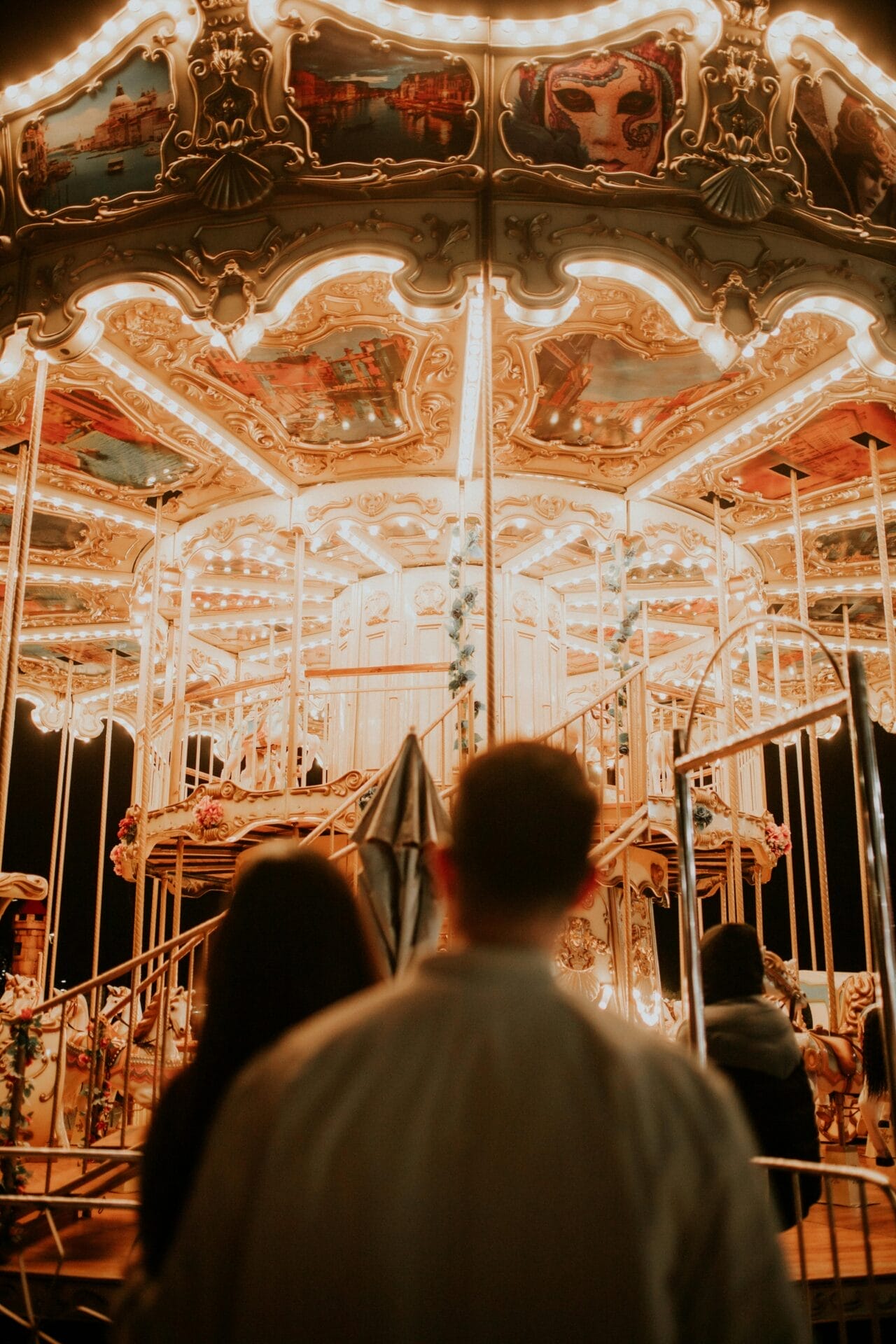 Un couple se tient devant un carrousel illuminé la nuit, rappelant un moment de rencontre parfait. Le carrousel, orné de lumières vives et de scènes peintes, crée une atmosphère chaleureuse et festive qui ne manquera pas de captiver même l'énigmatique homme Scorpion.