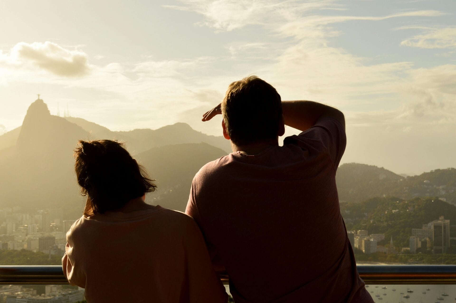 Deux personnes se tiennent sur un balcon surplombant une ville avec des montagnes en arrière-plan. Sous le chaud soleil de fin d'après-midi, l'homme Gémeaux se protège les yeux d'une main tandis que l'autre regarde au loin, réfléchissant aux complexités de la vie et aux aventures romantiques.