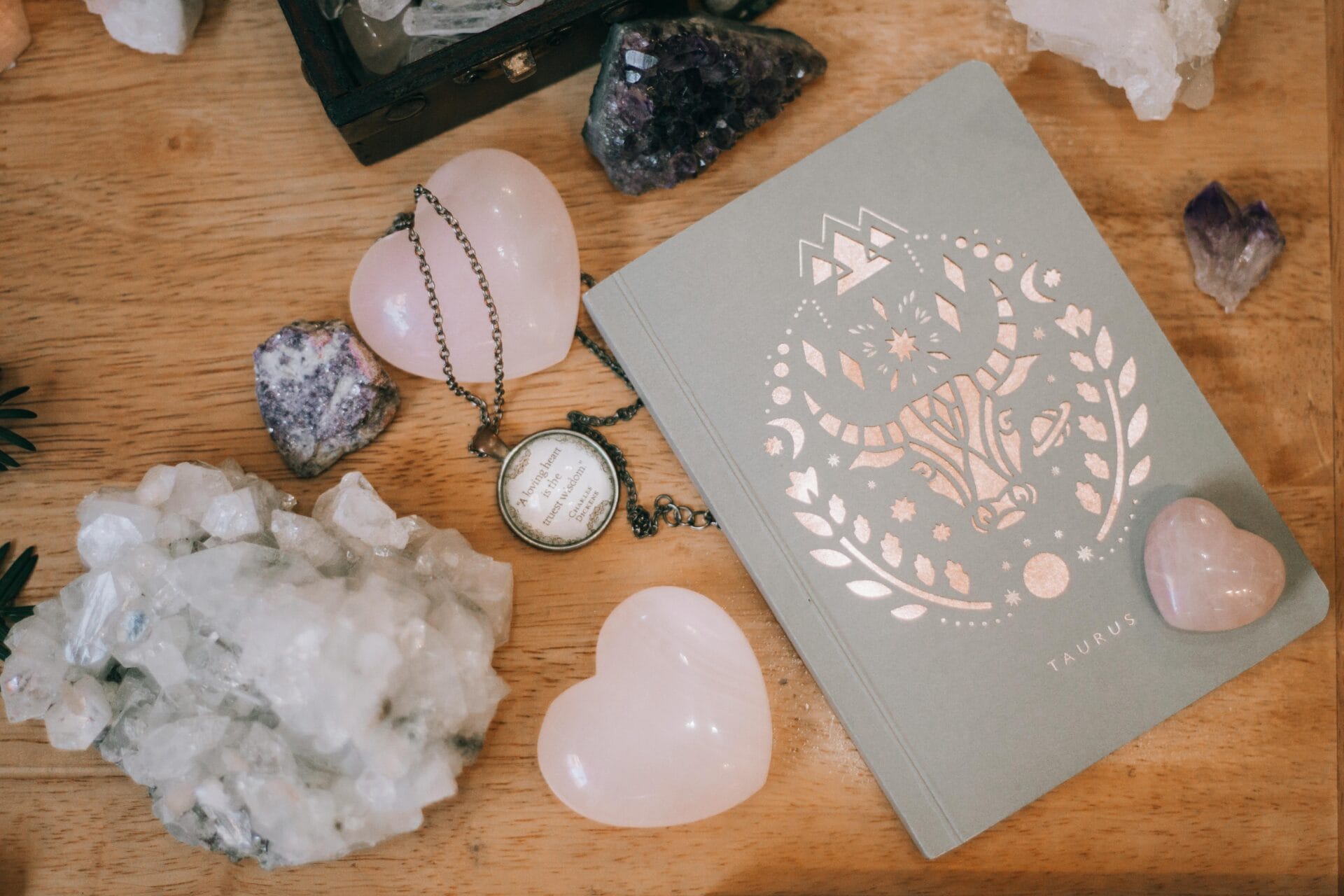 Une table en bois présente un journal sur le thème du Taureau avec un emblème du zodiaque, entouré de cristaux tels que des cœurs en quartz rose et des grappes d'améthyste. Un collier de montre à gousset de style ancien est posé à proximité, tandis que des conseils de rencontres pour un homme Scorpion attendent d'être découverts dans les pages du journal.