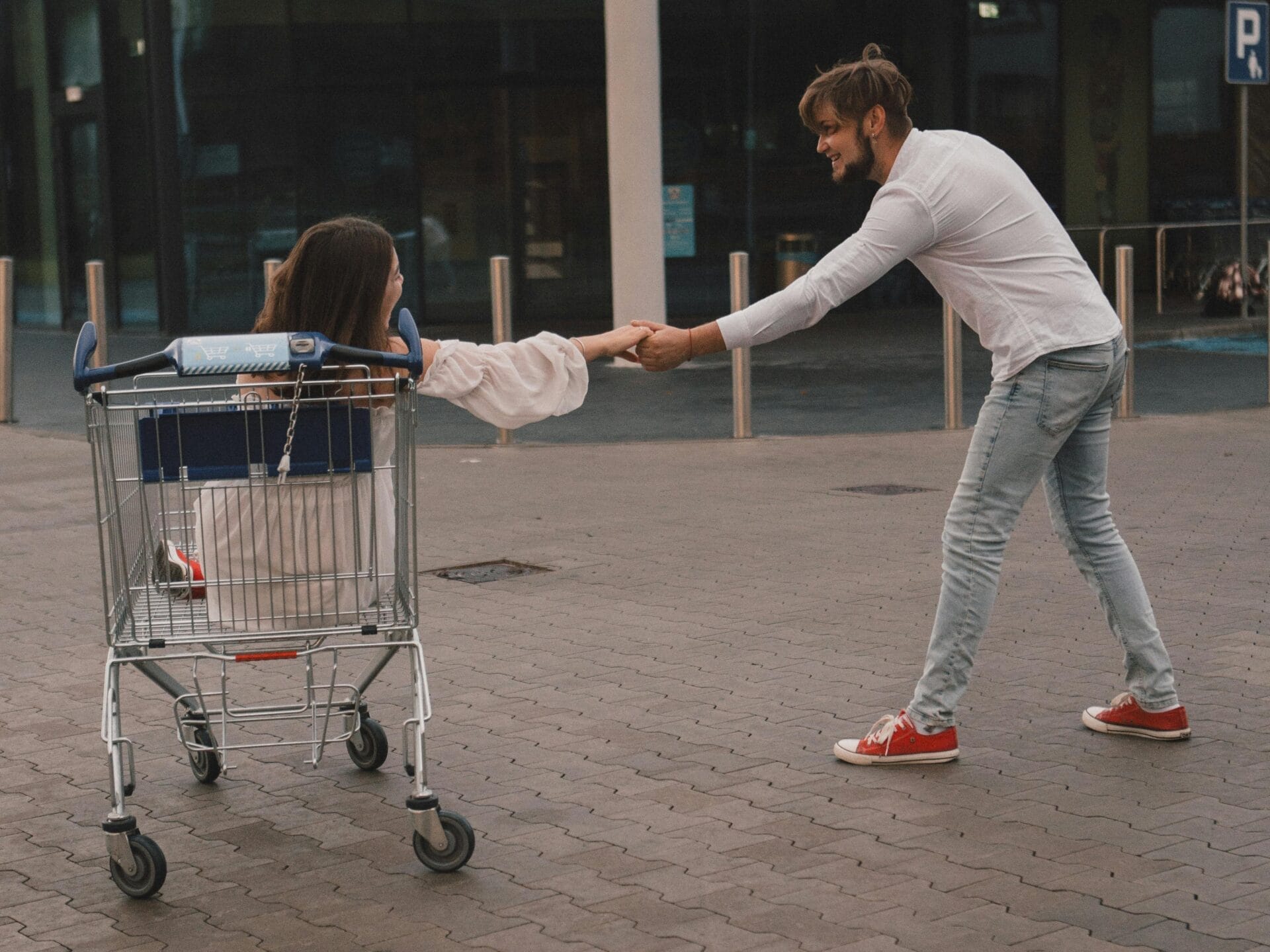 Une idée de rendez-vous amusante : un jeune homme tire joyeusement un chariot de supermarché avec une femme assise à l'intérieur, tous deux arborant un grand sourire. Il porte une chemise blanche et un jean avec des baskets rouges, tandis qu'elle est vêtue de blanc. À l'extérieur d'un bâtiment, sur une allée pavée, ils découvrent l'une des meilleures idées de rendez-vous gratuit : s'amuser ensemble de manière spontanée.