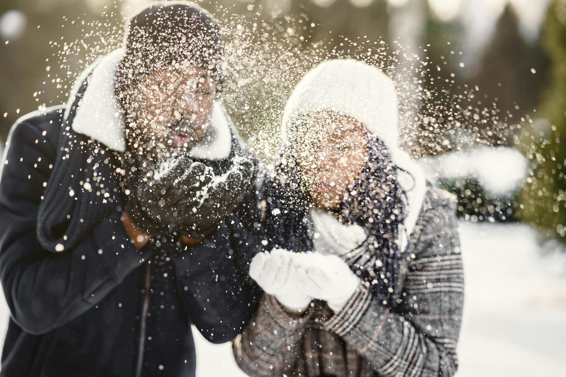 Deux personnes, emmitouflées dans des vêtements d'hiver, s'amusent à souffler de la neige de leurs mains, créant une gerbe étincelante. C'est une scène amusante qui se déroule au milieu de paysages et d'arbres enneigés, avec la lumière du soleil qui filtre à travers - une idée parfaite pour un premier rendez-vous gratuit pour ceux qui aiment la magie de l'hiver.