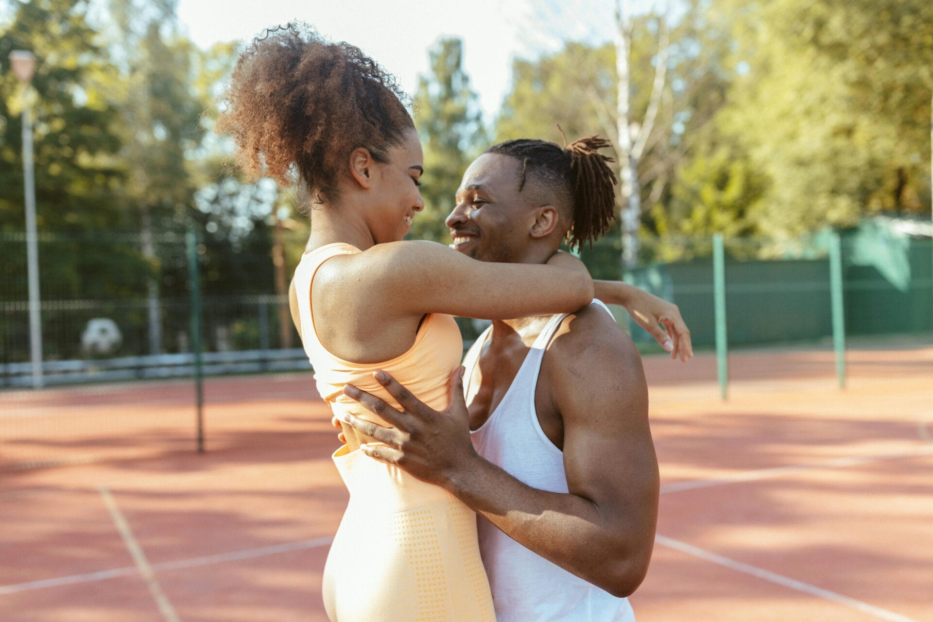 Un couple en tenue de sport s'étreint et sourit sur un court de tennis extérieur ensoleillé, profitant d'un moment de convivialité sobre et très apprécié. 