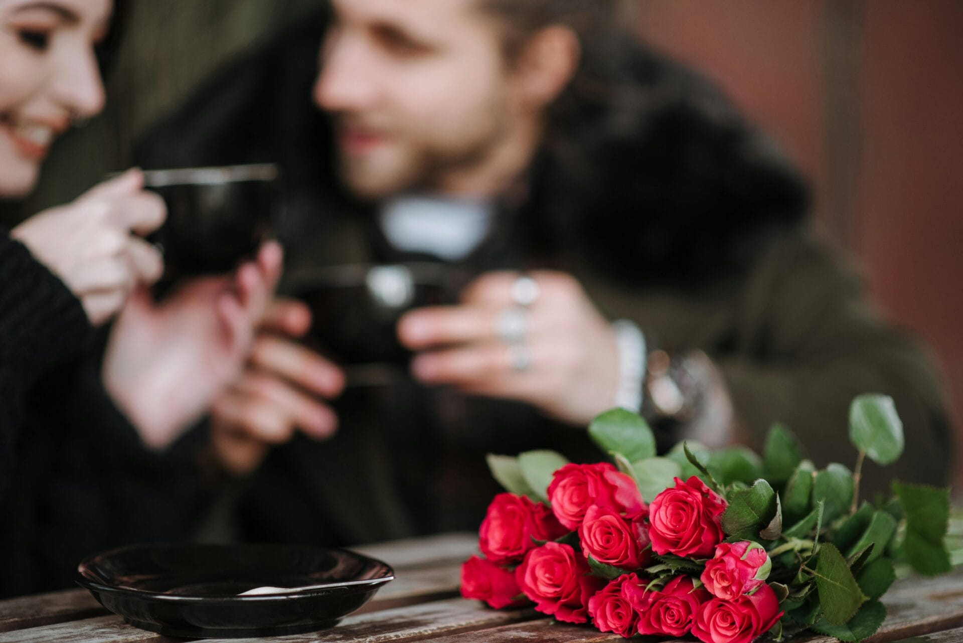 Un couple déguste une boisson chaude lors d'un rendez-vous sans alcool. Au premier plan, un bouquet de roses d'un rouge éclatant est posé à côté de tasses et de soucoupes noires, capturant l'essence de cette tendance populaire de la romance moderne.