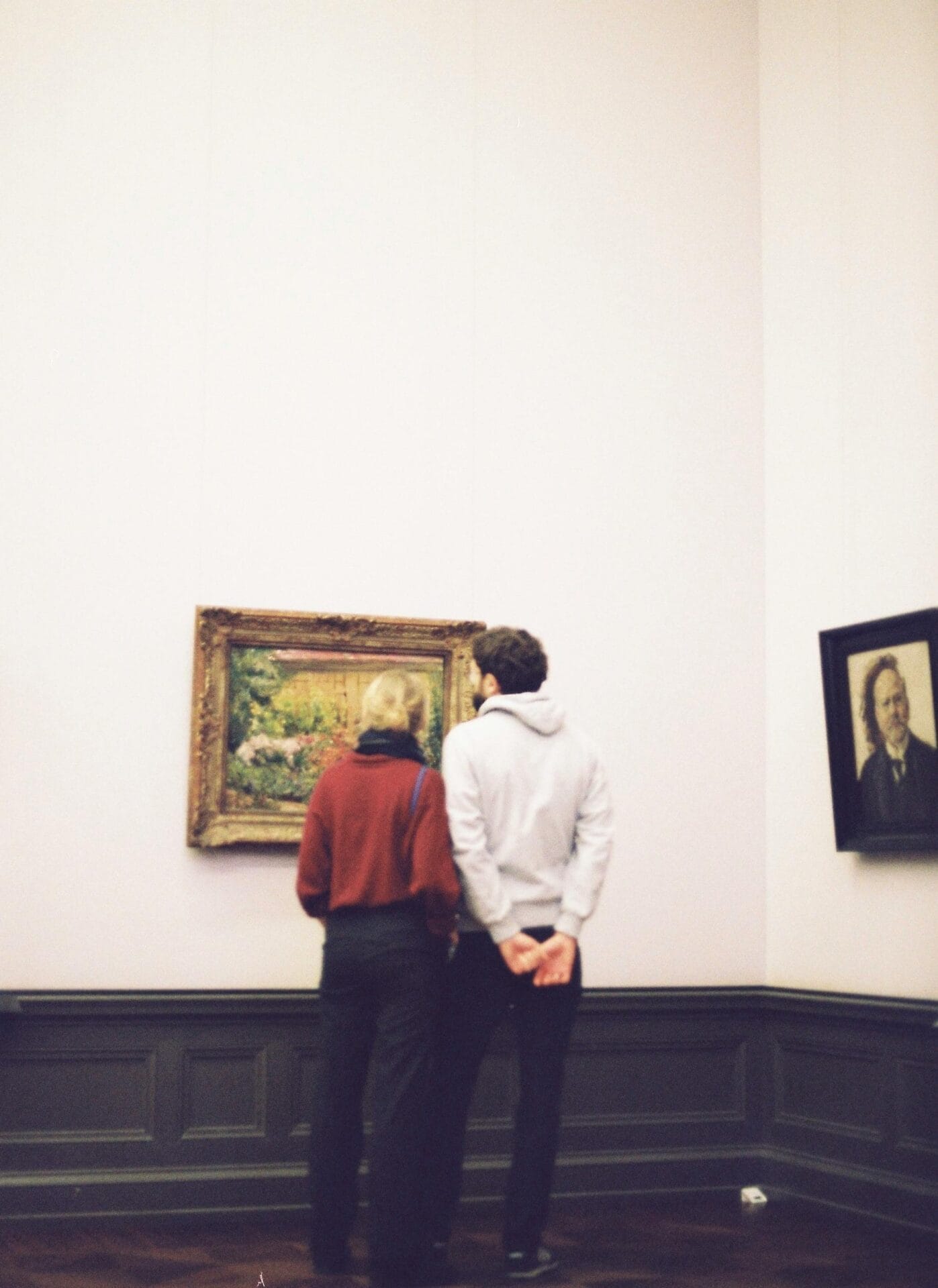 Deux personnes se tiennent côte à côte dans une galerie d'art, peut-être en rendez-vous, et regardent un tableau encadré. Ils sont dos à la caméra. La pièce a des murs crème et des boiseries foncées. Un autre portrait encadré est accroché au mur de droite, créant une atmosphère propice à ceux qui espèrent trouver l'amour en partageant des intérêts communs.