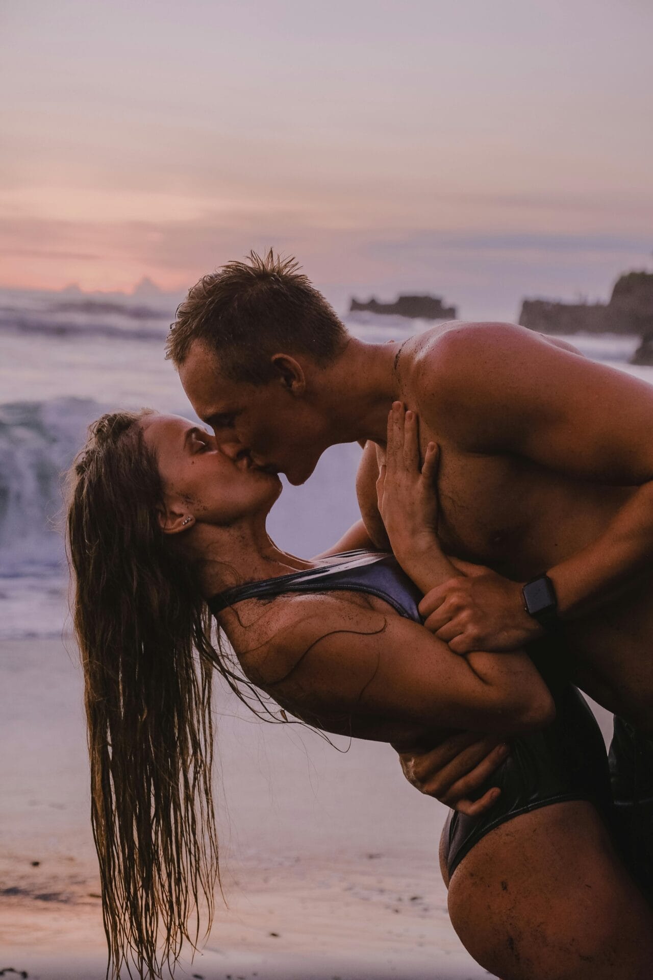 Un couple en maillot de bain s'embrasse passionnément sur une plage au coucher du soleil, incarnant l'attrait magnétique de la rencontre avec un homme Scorpion. Les vagues s'écrasent en arrière-plan et le ciel est peint dans des tons pastel. Ils sont tous les deux ensablés, l'air heureux et détendu au milieu du spectacle vibrant de la nature.