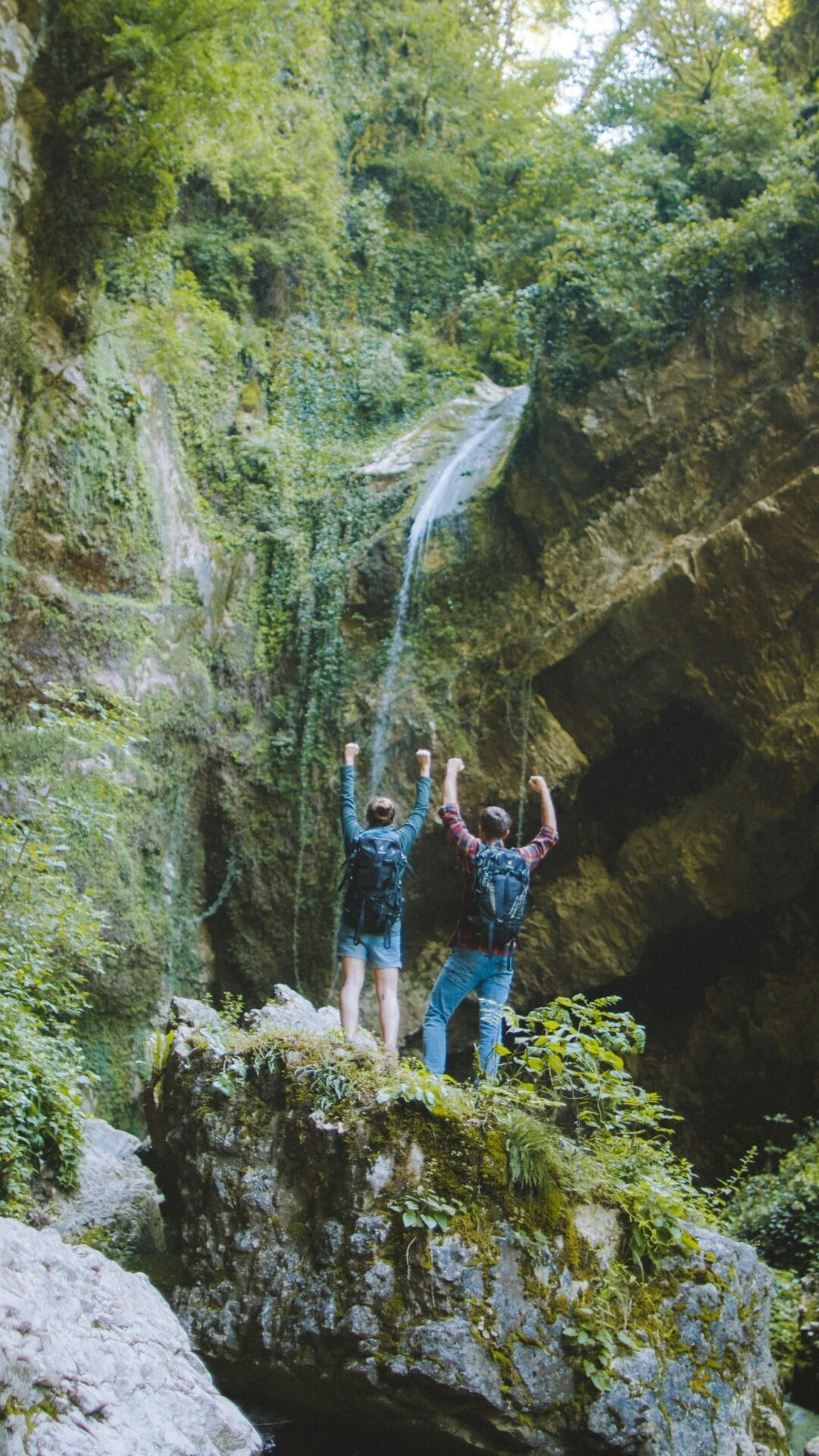 Deux personnes se tiennent triomphalement sur une corniche rocheuse, les bras levés, face à une cascade qui se jette dans une falaise verdoyante. Tels des jumeaux gémeaux fêtant une aventure commune, ils portent tous deux un sac à dos et semblent fêter leur arrivée dans ce lieu pittoresque.