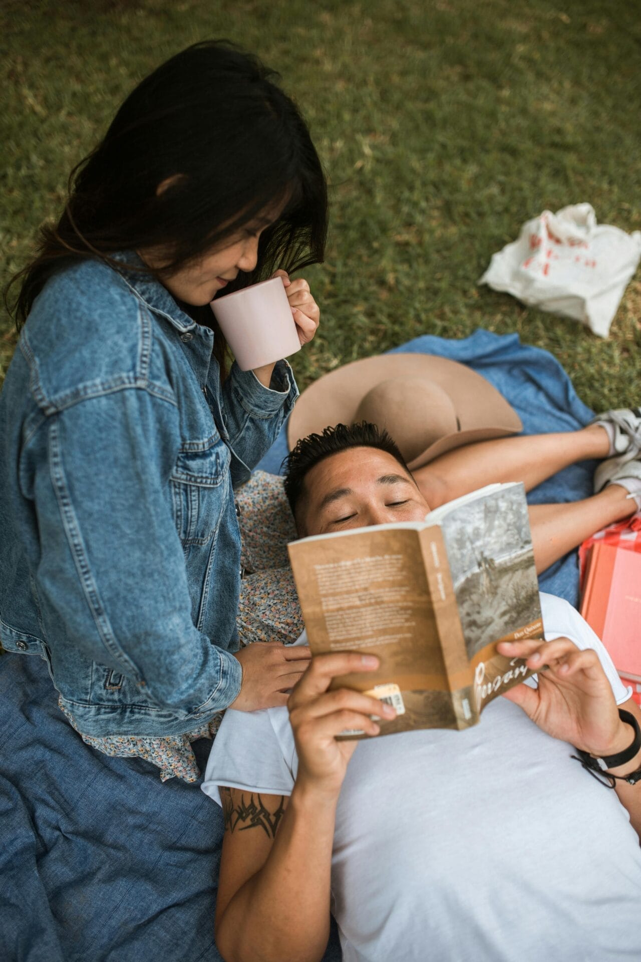 Une femme vêtue d'une veste en jean boit dans une tasse rose en se penchant sur son partenaire, qui est allongé sur une couverture et lit un livre. Ils profitent de l'extérieur sur l'herbe, trouvant l'amour dans un cadre paisible, avec un chapeau et un sac à côté d'eux.