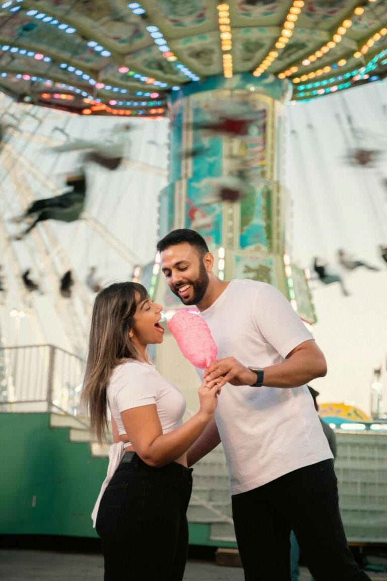 Lors de leur premier rendez-vous, le couple déguste une grande barbe à papa rose dans un parc d'attractions. Partageant un moment de jeu devant la balançoire décorée de lumières colorées, tous deux sourient dans leurs hauts blancs et leurs jeans décontractés.