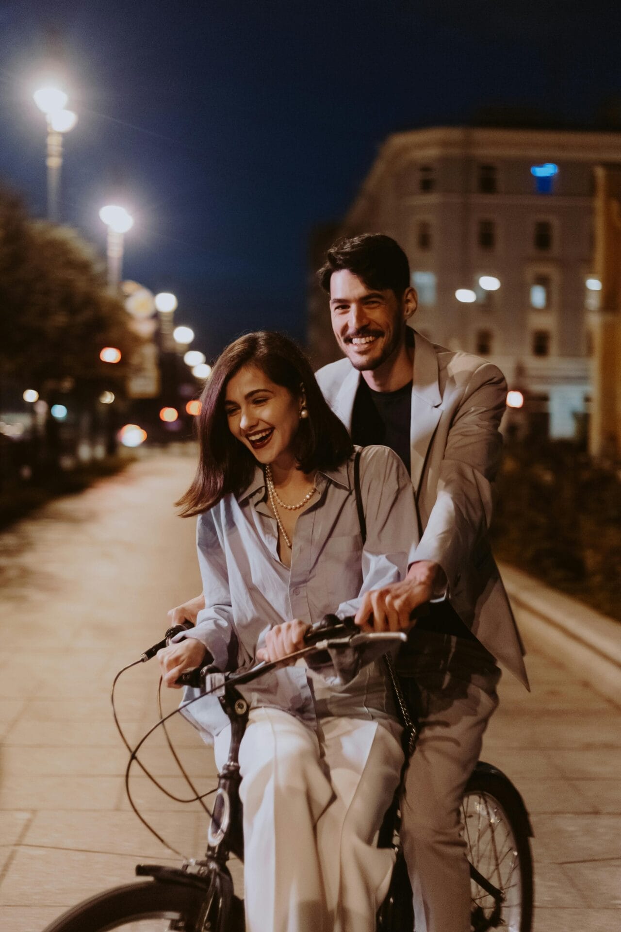 Une femme souriante fait du vélo la nuit et un homme souriant se tient à l'arrière, tenant le siège. Ils sont vêtus de tenues décontractées et élégantes, incarnant la joie des rencontres, alors qu'ils roulent dans les rues éclairées par les lampadaires, créant une atmosphère chaleureuse et animée dans leur quête de l'amour.