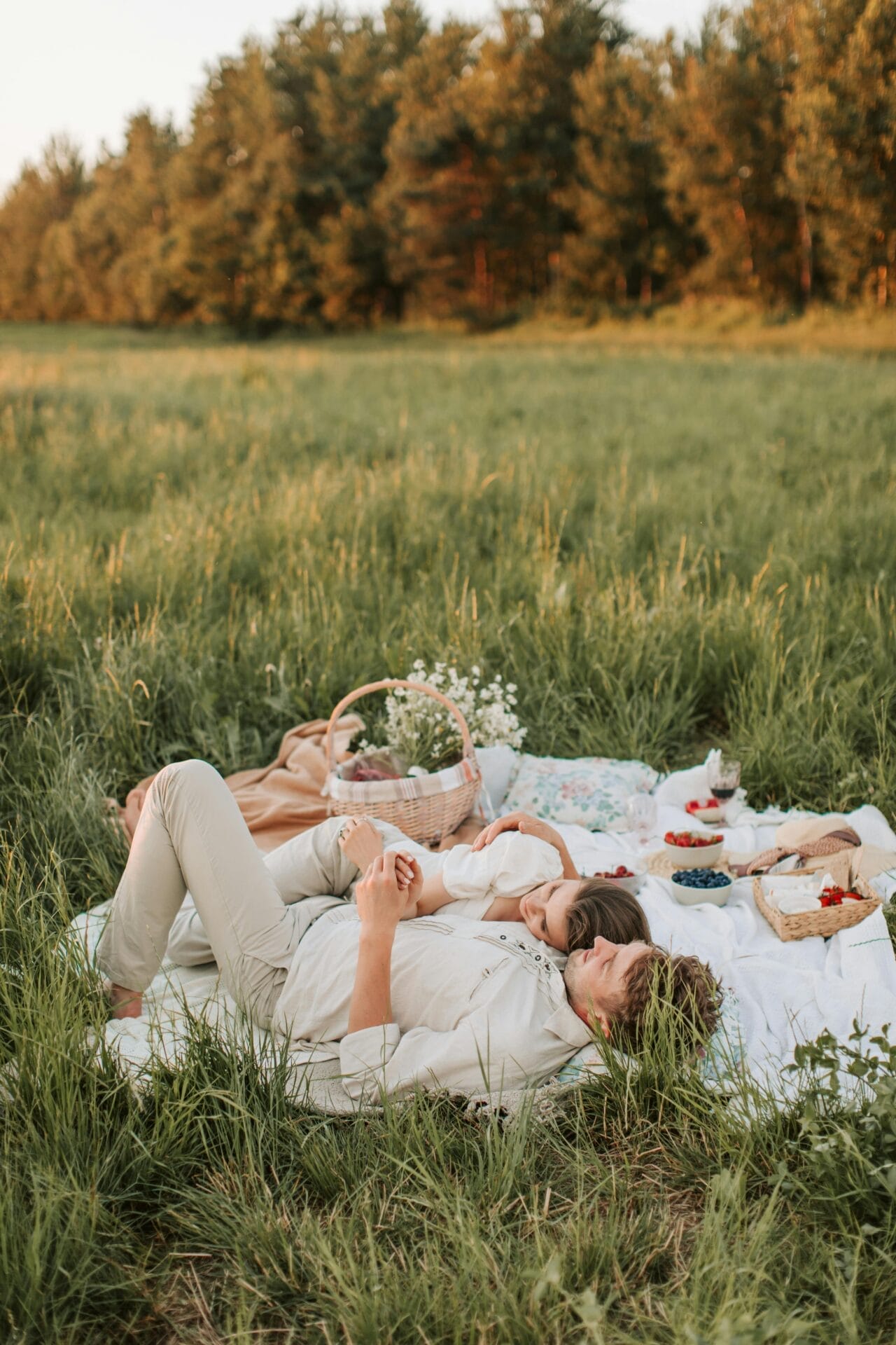 Une idée de rendez-vous : un couple profite d'un rendez-vous décontracté sur une couverture de pique-nique dans un champ herbeux, entouré d'une corbeille, de fleurs et d'assiettes de fruits. Détendus et satisfaits sous le ciel clair et les arbres en arrière-plan, c'est l'une des idées parfaites pour un premier rendez-vous.