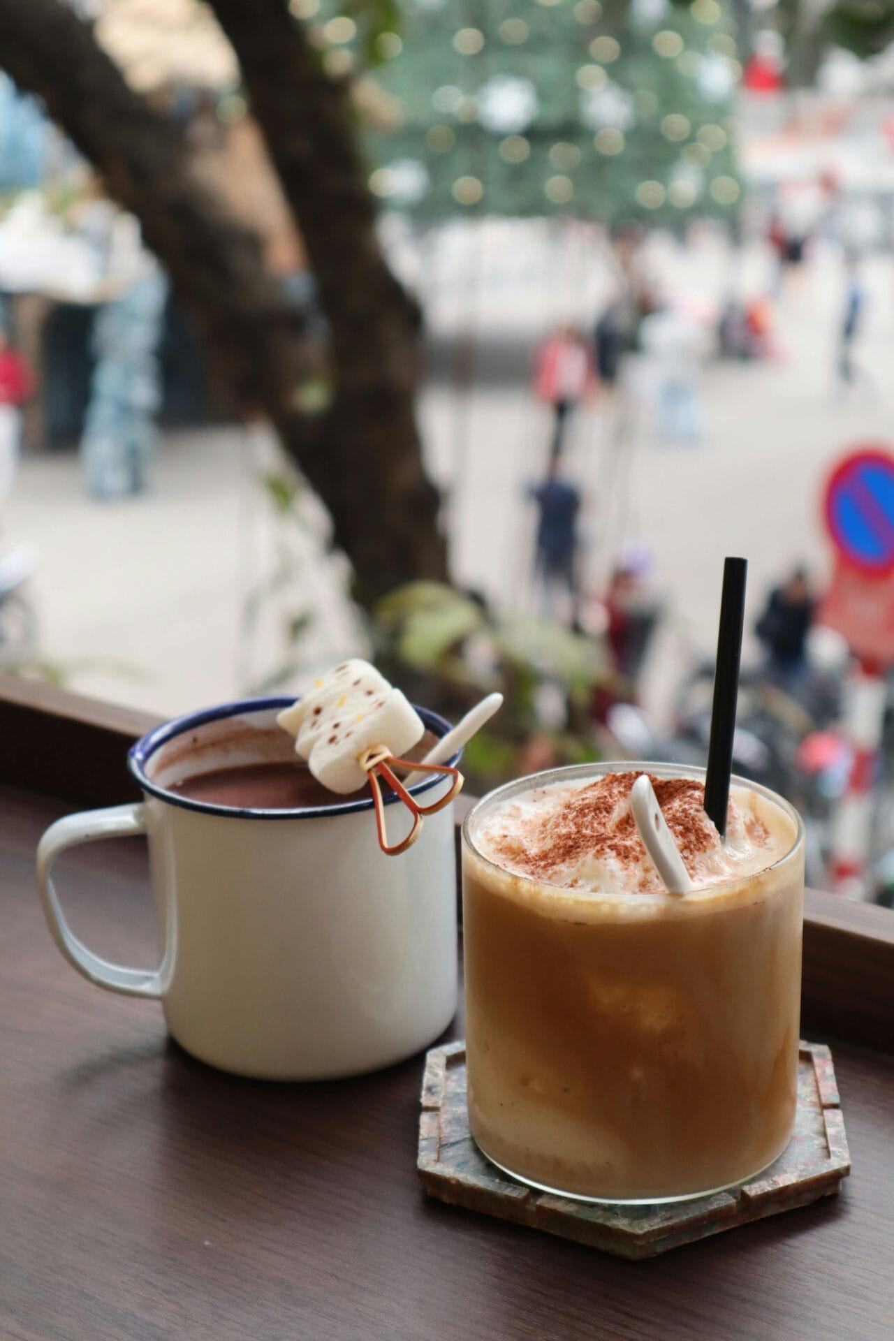 Une tasse de chocolat chaud avec des guimauves sur un bâton et un verre de café glacé avec de la crème et de la cannelle sont posés sur une table en bois. En arrière-plan, la scène urbaine floue reflète la tendance des rencontres sobres.