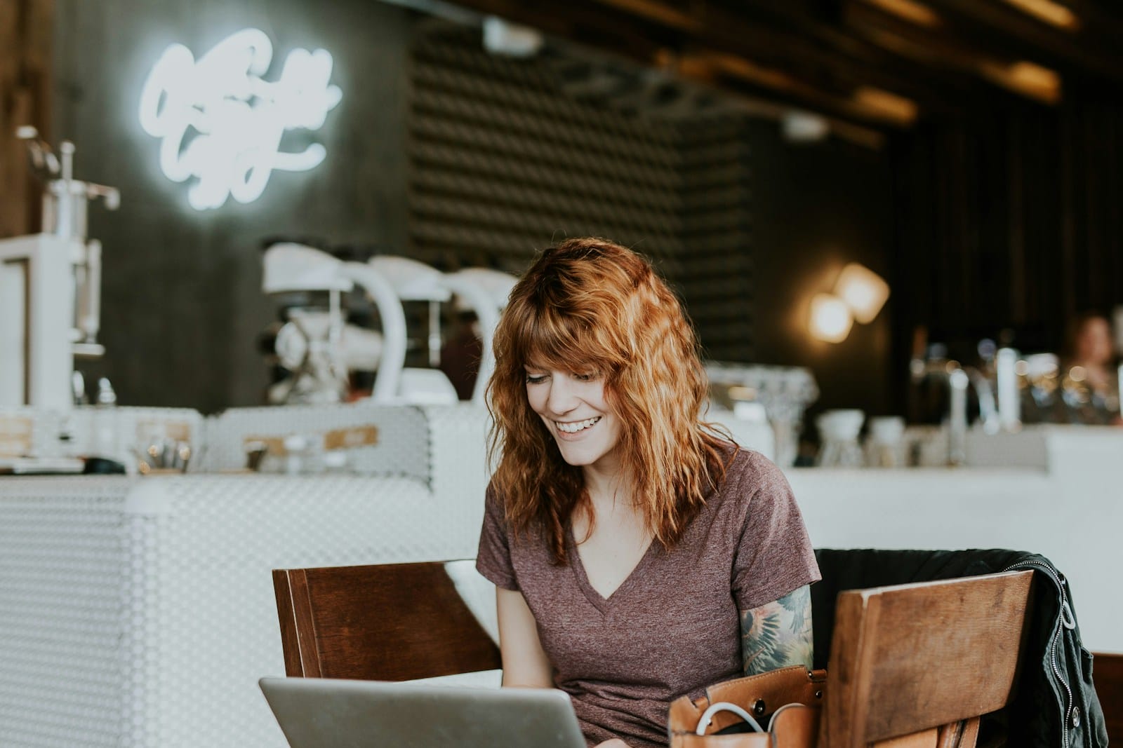 Une femme aux cheveux bruns ondulés est assise à une table dans un café, souriante, tandis qu'elle consulte son profil de rencontre en ligne sur son ordinateur portable. L'arrière-plan est composé d'une enseigne au néon floue et de machines à café. Elle porte un t-shirt marron et a un tatouage sur le bras gauche.