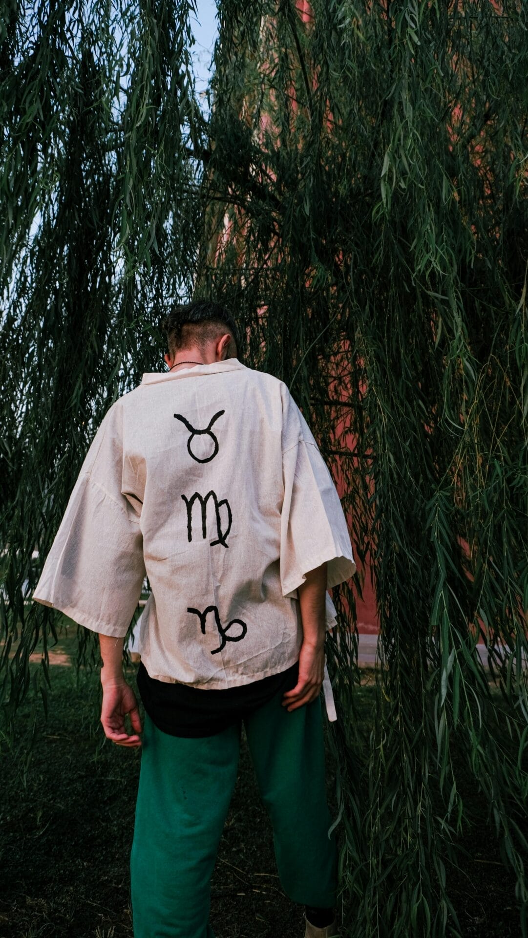 Une personne se tient sous des branches d'arbre tombantes, vêtue d'une chemise beige avec des symboles noirs et d'un pantalon vert. Elle tourne le dos à l'appareil photo, réfléchissant peut-être à la compatibilité avec l'homme Taureau. L'arrière-plan comprend une végétation luxuriante et une partie d'un bâtiment, créant ainsi une ambiance contemplative propice aux réflexions sur la compatibilité.